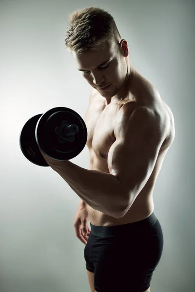 Pesos antes de las fechas. Estudio de un joven musculoso ejercitándose con una mancuerna sobre un fondo gris. —  Fotos de Stock