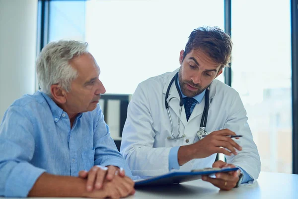 Elke patiënt moet een duidelijk begrip hebben van zijn diagnose. Schot van een knappe dokter die wat papierwerk doorneemt met een mannelijke senior patiënt in een kliniek. — Stockfoto