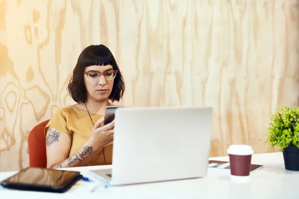 A enviar algumas mensagens a uns amigos do escritório. Tiro de uma jovem designer usando seu celular enquanto trabalhava em um laptop em um escritório. — Fotografia de Stock