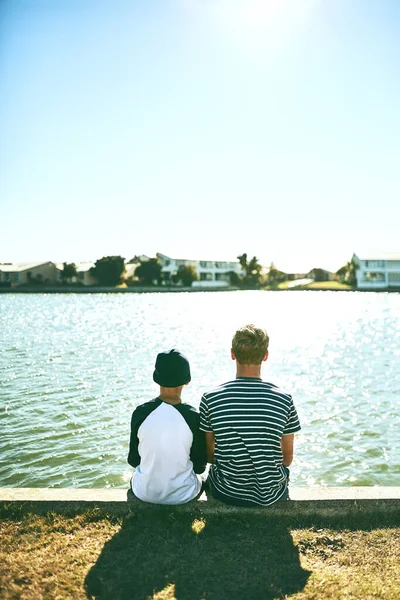 A apreciar a vista. Rearview tiro de dois irreconhecíveis jovens irmãos sentados do lado de fora por uma lagoa. — Fotografia de Stock