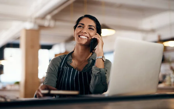 No es de extrañar que su café sea un gran éxito. Fotografía de una mujer joven usando un ordenador portátil y un teléfono inteligente mientras trabajaba en un café. — Foto de Stock