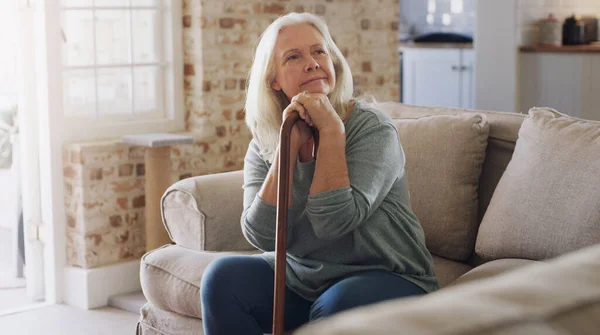 Me pregunto qué podría hacer hoy. Foto de una mujer mayor sentada sola en el sofá en casa y mirando contemplativa mientras sostiene su bastón. — Foto de Stock