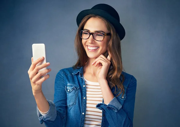 Questo cappello è degno di selfie. Girato di una giovane donna allegra che usa il suo cellulare su uno sfondo grigio in studio. — Foto Stock