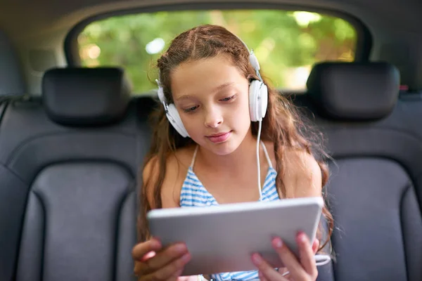 A ver os filmes dela no banco de trás. Tiro de uma jovem sentada em um banco de trás do carro usando fones de ouvido e usando um tablet digital. — Fotografia de Stock