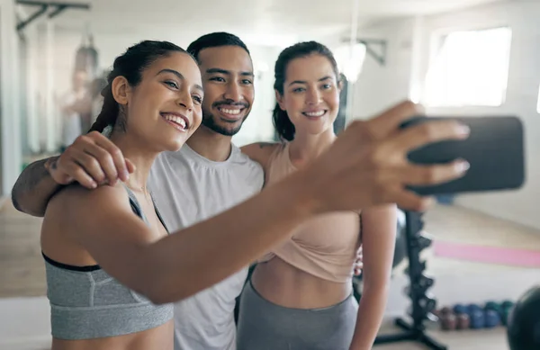 Online para espalhar alguma inspiração fitness. Tiro de três jovens atletas tirando uma selfie enquanto estavam juntos no ginásio. — Fotografia de Stock