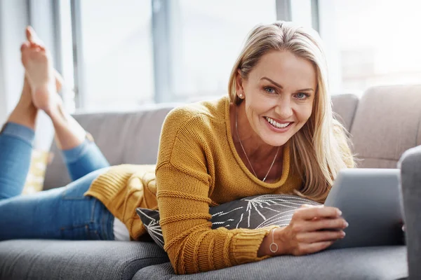 My go to gadget for downtime entertainment. Portrait of an attractive mature woman using a digital tablet while relaxing on the sofa at home. — Stock Photo, Image