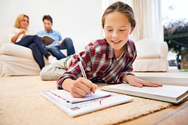 Homework done equals relaxing time. Shot of a little girl lying on the living room floor doing homework with her parents in the background. Stock Image