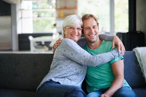 Me encanta que me lo ofrezca. Retrato de una mujer mayor abrazando a su hijo mientras está sentada en el sofá en casa. —  Fotos de Stock
