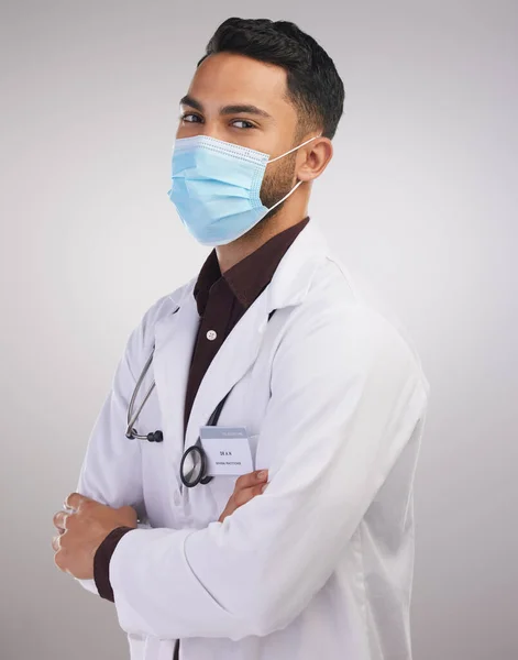 You better be following Covid rules. Shot of a handsome young doctor standing alone in the studio with his arms folded and wearing a face mask. — ストック写真