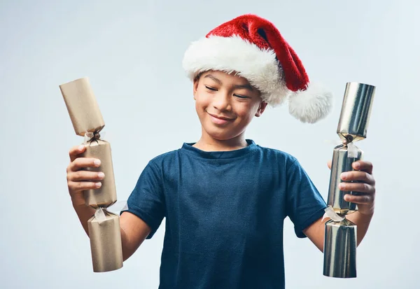 크리스마스 크래커 폭발이 기다리고 있습니다. 스튜디오는 산타 모자를 쓰고 회색 배경에 크리스마스 크래커 2 개를 들고 있는 귀여운 소년의 사진을 찍었습니다.. — 스톡 사진