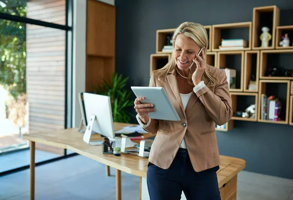 Fusionando tecnología con éxito. Fotografía de una mujer de negocios madura usando una tableta digital mientras habla en un teléfono celular en una oficina. —  Fotos de Stock