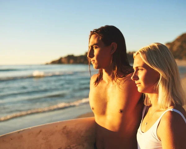Ψάχνω το τέλειο κύμα. Τραβηγμένο πλάνο ενός νεαρού ζευγαριού με μια σανίδα του σερφ να στέκεται μαζί και να βλέπει το ηλιοβασίλεμα στην παραλία. — Φωτογραφία Αρχείου