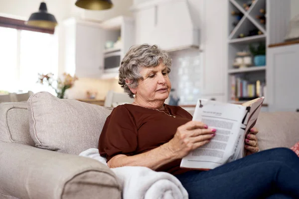 Capítulo escolhido de contentamento. Tiro de uma mulher idosa relaxante com um livro no sofá em casa. — Fotografia de Stock