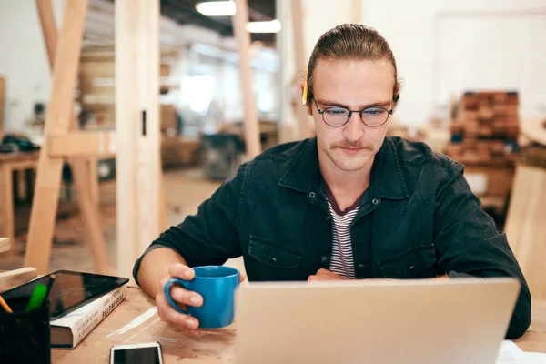 Estoy comprobando algunos pedidos de clientes en línea. Recortado disparo de un joven carpintero guapo bebiendo café y trabajando en un portátil dentro de su taller. — Foto de Stock