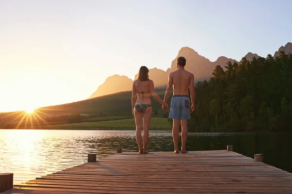 Zeit für einen Sonnenuntergang. Rückansicht eines jungen Paares in Badeanzügen, das bei Sonnenuntergang auf einer Anklagebank steht. — Stockfoto