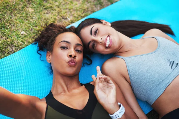 Última selfie para a estrada. Retrato recortado de duas jovens mulheres atraentes deitadas em um tapete de ioga e posando para uma selfie ao ar livre. — Fotografia de Stock