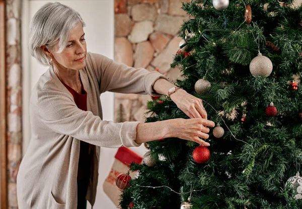 Podes ir por aqui. Cortada tiro de uma mulher madura despreocupada colocando decorações em uma árvore de Natal dentro de casa durante o dia. — Fotografia de Stock