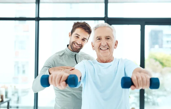 Er setzt sich für ein gesünderes Leben ein. Aufnahme eines freundlichen Physiotherapeuten, der seinem älteren Patienten beim Training mit Gewichten hilft. — Stockfoto