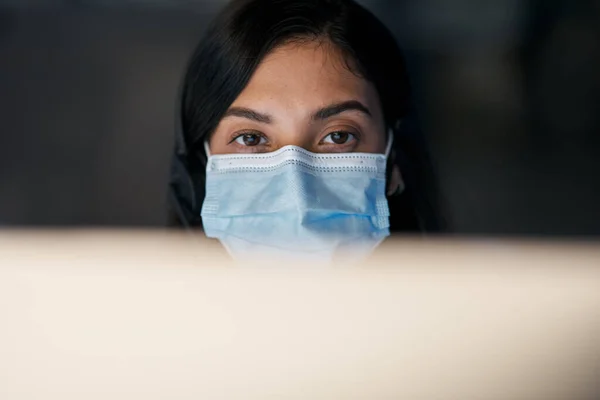 Os clientes vêm em primeiro lugar, mesmo em face de uma pandemia. Tiro de uma jovem mascarada usando um fone de ouvido e computador tarde da noite em um escritório moderno. — Fotografia de Stock