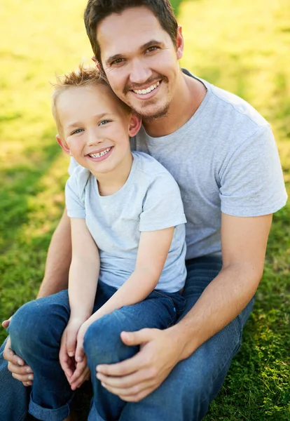 Улыбки и солнце в парке. Портрет улыбающегося отца и его сына, сидящего на траве в парке. — стоковое фото