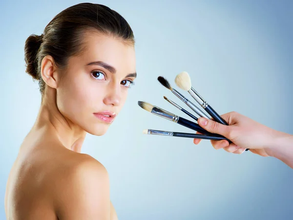 Make-up je můj výraz. Studio portrét ruky držící make-up štětce vedle krásné mladé ženy na modrém pozadí. — Stock fotografie