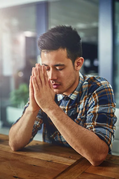 Seeking out some guidance and strength. Shot of a young designer sitting in an office with his hands clasped in prayer. — ストック写真