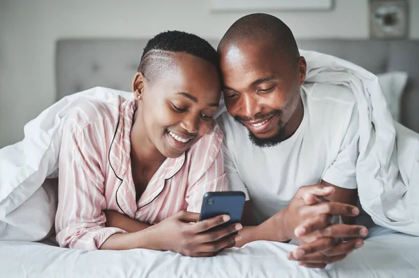 Hoje em dia, há mais de uma forma de nos ligarmos. Tiro de um jovem casal feliz usando um smartphone juntos no quarto em casa. — Fotografia de Stock