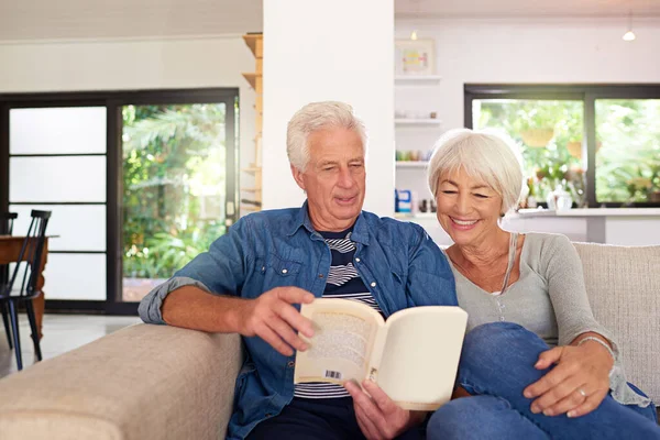 Entre as páginas de um livro de amor. Tiro de um casal de idosos relaxados lendo um livro juntos no sofá em casa. — Fotografia de Stock