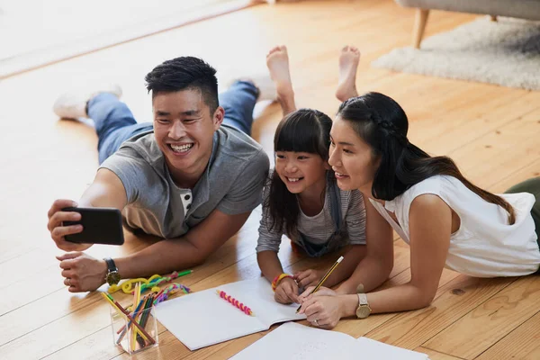 Pre homework selfie. Shot of a cheerful mother and father doing homework with their little girl while taking a self portrait at home during the day. — Stock Photo, Image