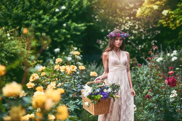 彼女のバスケットは品種で咲いています。花頭の花輪を身に着けている美しい若い女性のショットと自然の中で花でいっぱいのバスケットを保持. — ストック写真