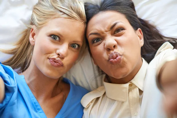Haz una cara. Retrato de ángulo alto de dos mujeres jóvenes atractivas tomando selfies mientras están acostadas en una cama. —  Fotos de Stock