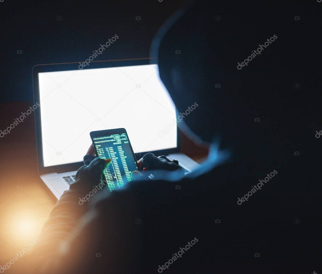 Gaining access to all your passwords. Shot of an unrecognisable hacker using a cellphone and laptop in the dark.