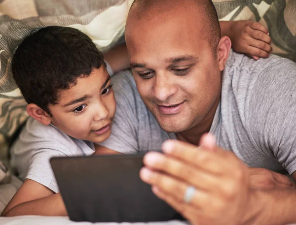 Ze lijken te genieten van hun show samen. Gehakte foto van een vrolijke jongeman en zijn zoon samen video 's aan het bekijken op een digitale tablet terwijl ze overdag thuis op een bed rondhangen. — Stockfoto