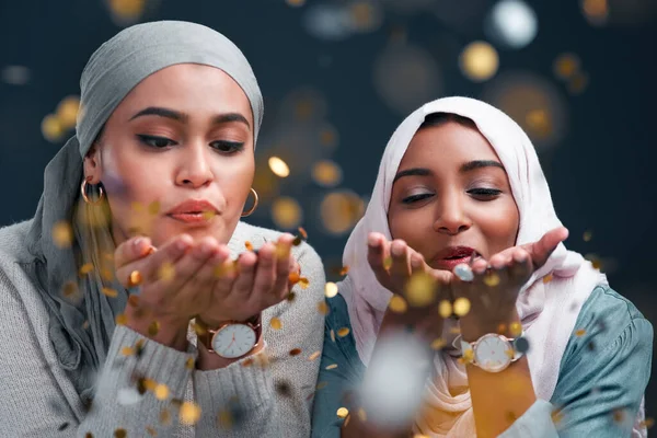 Leave a little sparkle everywhere you go. Cropped shot of two attractive young women wearing hijabs and blowing confetti while standing against a black studio background. — ストック写真