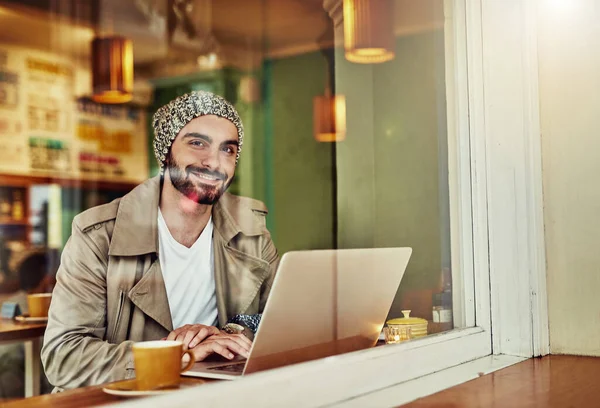 Nongkrong di kafe favoritnya. Potret seorang pemuda bergaya tersenyum sambil minum kopi dan menggunakan laptop di sebuah kafe. — Stok Foto