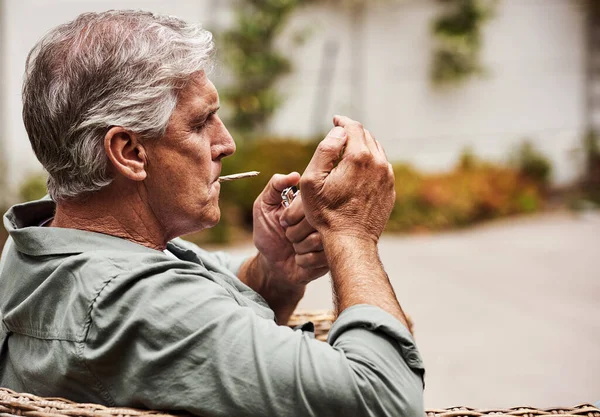 Hora de iluminar. Una foto de un anciano relajado fumando un porro de marihuana solo dentro de su jardín en casa durante el día. —  Fotos de Stock