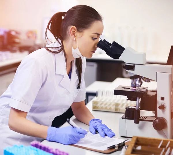 A verdade está debaixo da lente. Tiro de um jovem cientista tomando notas ao usar um microscópio em um laboratório. — Fotografia de Stock