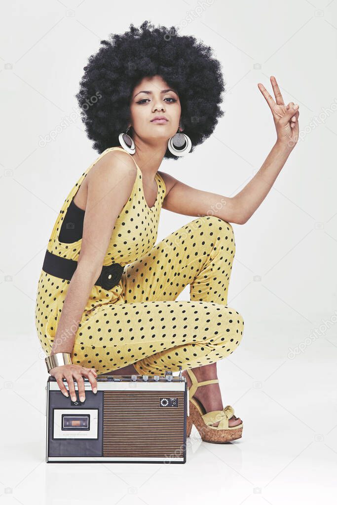 That 70s attitude.... A young woman wearing a 70s retro jumpsuit sitting by a cassette player showing the peace sign.