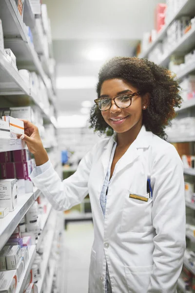 Тобі щось потрібно, у нас є. Портрет веселого молодого жіночого фармацевта, який кладе ліки на полиці, дивлячись на камеру в аптеці . — стокове фото