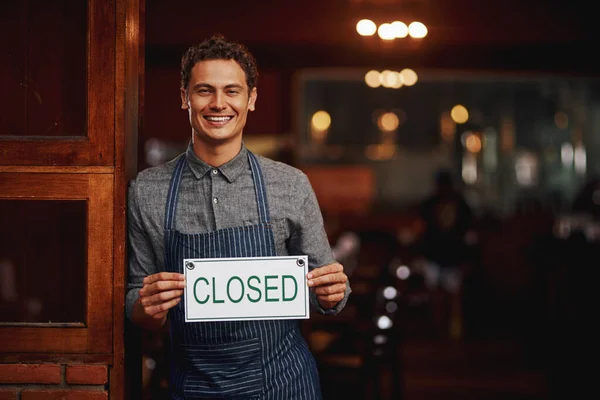 Sorry, kom een andere keer terug. Portret van een vrolijke jonge ondernemer die een bord omhoog houdt waarop staat gesloten terwijl hij overdag onder een deuropening bij een bierbrouwerij staat. — Stockfoto