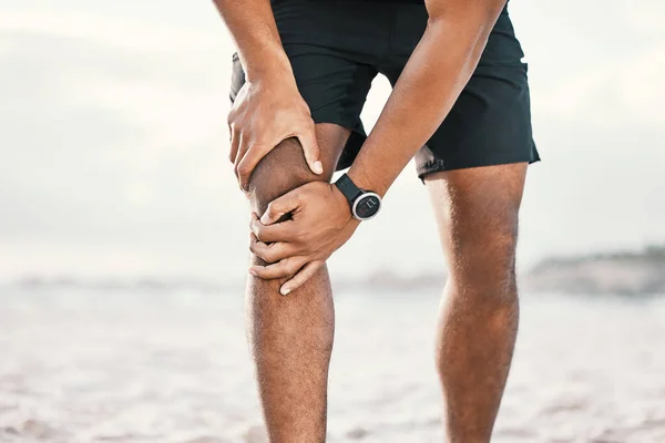 Parece que no más correr. Cortada la inyección de un atleta masculino irreconocible sosteniendo su rodilla con dolor mientras hace ejercicio en la playa. —  Fotos de Stock