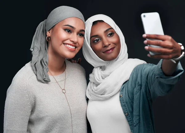 Selfie-Zeit. Schnappschuss von zwei attraktiven jungen Frauen, die zusammen stehen und Hijabs tragen, während sie ein Selfie vor schwarzem Hintergrund machen. — Stockfoto