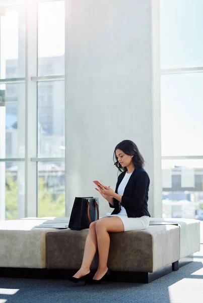 Taking time to reply to her clients. Full length shot of an attractive young businesswoman using a smartphone while sitting in a waiting room. — ストック写真