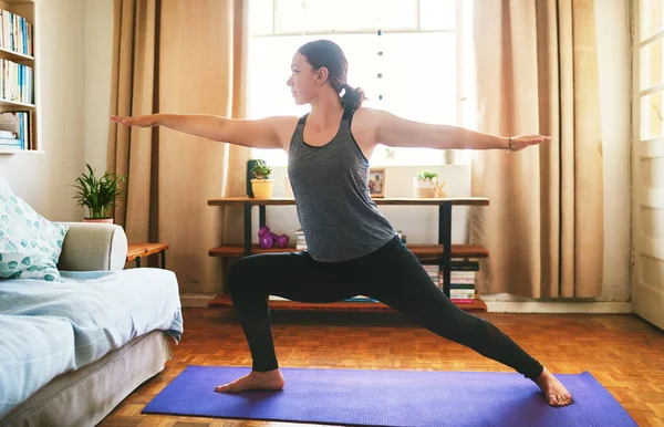 Siente la quemadura. Foto recortada de una atractiva joven de pie y sosteniendo una pose de guerrera mientras hace yoga en su casa. — Foto de Stock
