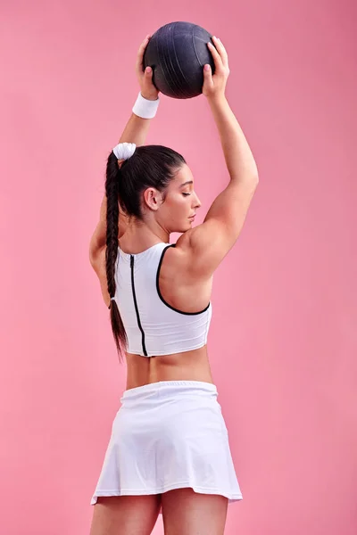 Stående lång och stark. Studio skott av en sportig ung kvinna som håller en boll över huvudet mot en rosa bakgrund. — Stockfoto