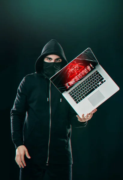 Tu pourrais être le prochain. Portrait d'un pirate informatique équilibrant un ordinateur portable tout en se tenant debout sur un fond sombre. — Photo