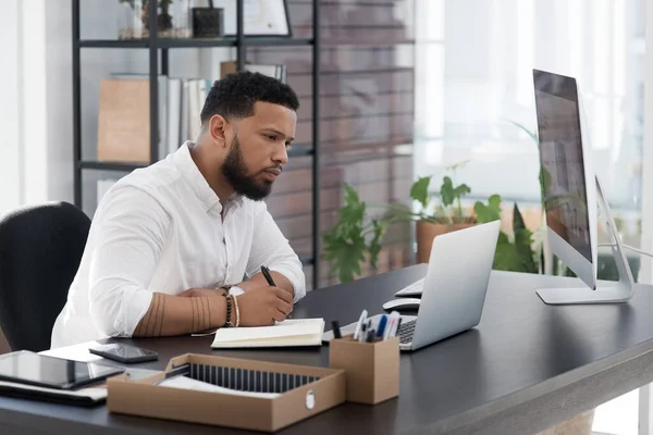 Sitting down for some thorough planning. Shot of a young businessman writing notes while working on a laptop in an office. — Stock Photo, Image