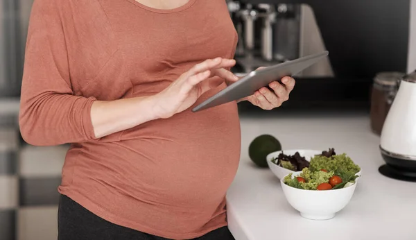 Vyhledávám nějaké recepty online. Vystřižený záběr těhotné ženy pomocí svého tabletu při jídle salátu v kuchyni. — Stock fotografie