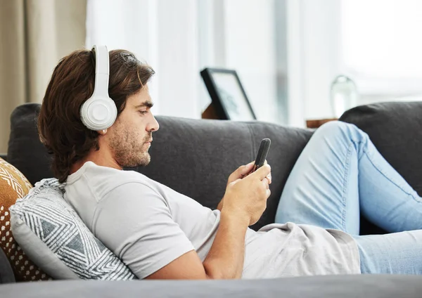 Sending a risky text has never been this nerve wracking. Shot of a young man relaxing on his sofa in the lounge while using his phone and headphones. — Stock Photo, Image