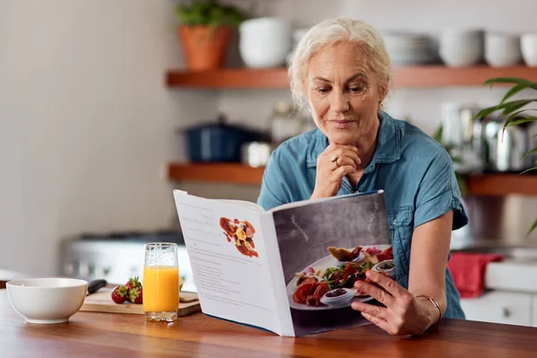 A maneira saudável é a maneira inteligente. Tiro de uma mulher madura lendo um livro enquanto prepara o café da manhã em casa. — Fotografia de Stock